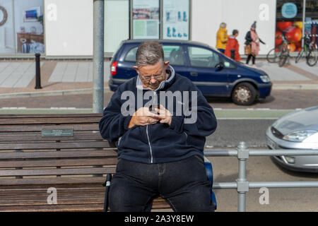 A middle aged overweight man sitting on a bench looking at his mobile phone or cell phone Stock Photo