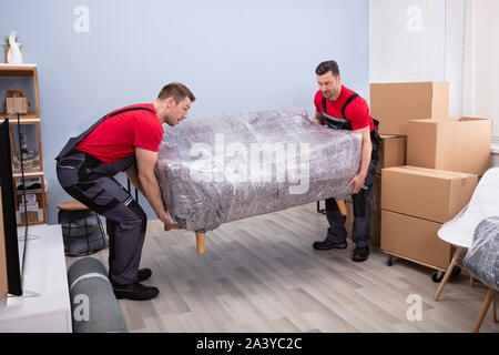 Portrait Of Two Young Happy Male Movers Carrying Wrapped Sofa In New House Stock Photo
