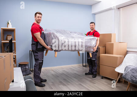 Portrait Of Two Young Happy Male Movers Carrying Wrapped Sofa In New House Stock Photo