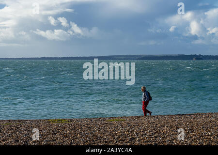 A middle aged woman wearing a backpack walking along a pebble beach with the sea in the background Stock Photo