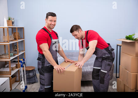 Portrait Of Two Smiling Young Male Movers In Uniform Packing Cardboard Boxes In Living Room Stock Photo