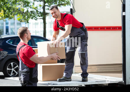 Two Young Male Movers In Uniform Carrying Cardboard Boxes From Truck Stock Photo
