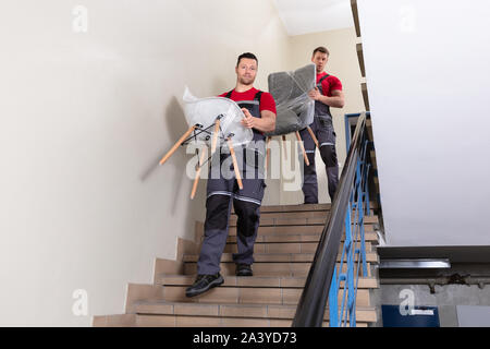 Portrait Of A Young Male Movers In Uniform Carrying Chairs Walking Downward On Staircase Stock Photo