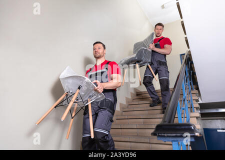 Portrait Of A Young Male Movers In Uniform Carrying Chairs Walking Downward On Staircase Stock Photo
