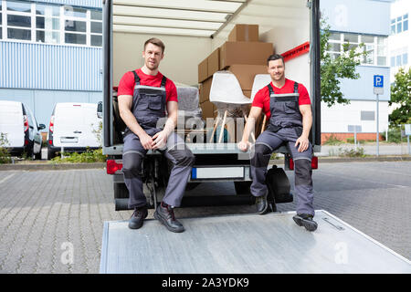 Portrait Of Smiling Movers Sitting In Van With Stack Of Cardboard Boxes And Furniture Stock Photo