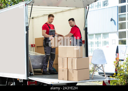 Two Young Men Unloading And Stacking The Brown Cardboard Boxes On Moving Truck Stock Photo