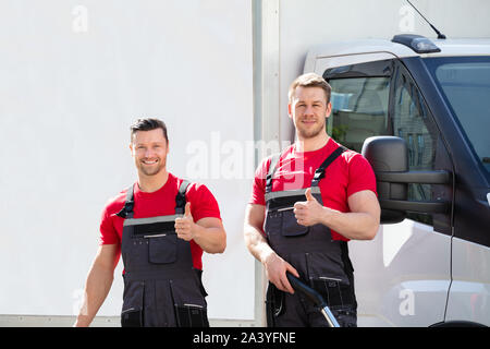 Portrait Of Two Male Janitors In Uniform Showing Thumbs Up Sign Standing Against Parked Truck Stock Photo