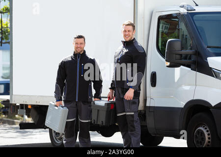 Portrait Of Smiling Two Male Electrician Carrying Tool Boxes Standing Against Truck Looking At Camera Stock Photo