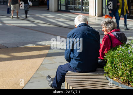 An elderly couple sat on a bench talking at a shopping mall or shopping centre Stock Photo