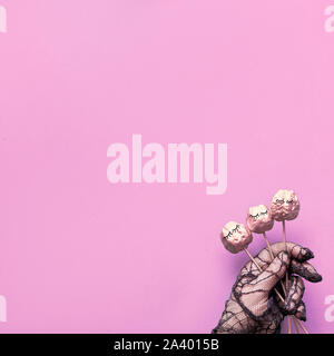 Creative Halloween flat lay on pink paper background, square with copy-space. Hand in mesh glove holding small pumpkins on sticks with closed eyes. Stock Photo