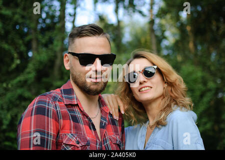 Lovely couple of bearded hipster boyfriend in red shirt and blond girlfriend in blue casual jeans blouse looking at the camera wearing dark sunglasses Stock Photo
