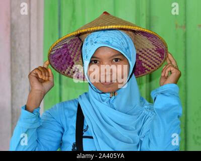 Young Indonesian Muslim woman wears on top of her turquoise hijab an Asian conical straw hat (caping) with built-in head support.