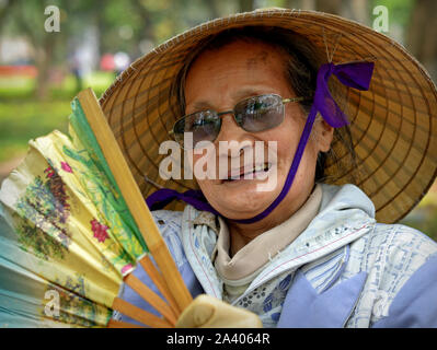 Elderly Vietnamese woman with Asian conical hat poses for the camera with a traditional handheld bamboo fan. Stock Photo