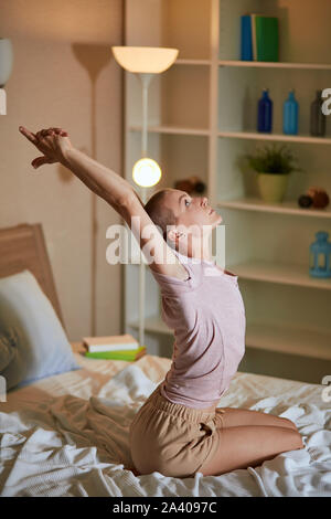 Pretty caucasian woman sit and practice at home with raised arms, yoga in everyday life. Yoga every morning. Flexible short-haired girl on bed Stock Photo