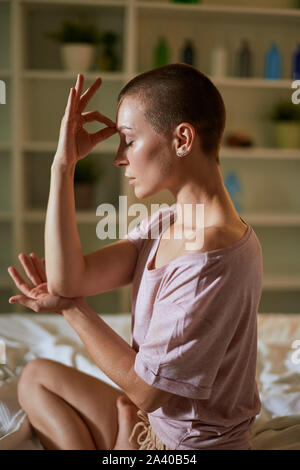 Relaxing woman practice yoga exercises at home on bed. Fit female in pajamas sits with crossed legs on white bed. Background bedroom Stock Photo