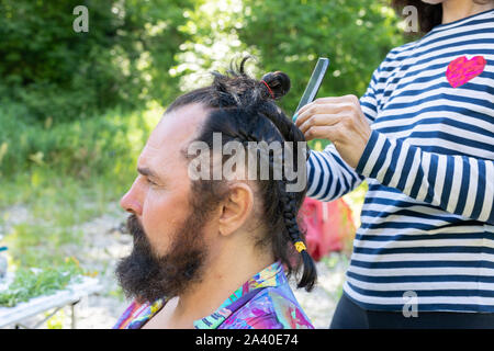 doing a haircut for men with long black hair. girl braids a pigtail to a middle-aged bearded man Stock Photo