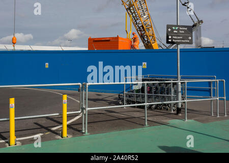A construction crane and workman appear over airport baggage trolleys and blue hoarding at City Airport, on 10th October 2019, east London, England. Stock Photo