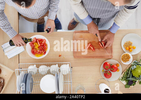 High angle view of two men standing at the kitchen table and preparing vegetable salad together in the kitchen Stock Photo