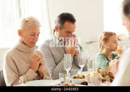 Family of three sitting at the table and praying together before the dinner in domestic room Stock Photo