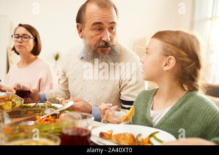 Grandfather talking to his granddaughter while they have dinner at the table with other members of the family at home Stock Photo