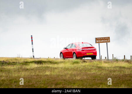 A red car passing a sign reading, Thanks You For visiting Weardale, on a moorland road in County Durham, England, UK Stock Photo
