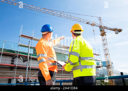 Rear View Of Two Male Architect Wearing Hardhat Looking At Construction Site Stock Photo