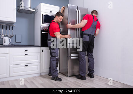 Two Young Male Movers In Uniform Placing Modern Steel Refrigerator In Kitchen Stock Photo