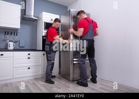 Two Young Male Movers In Uniform Placing Modern Steel Refrigerator In Kitchen Stock Photo