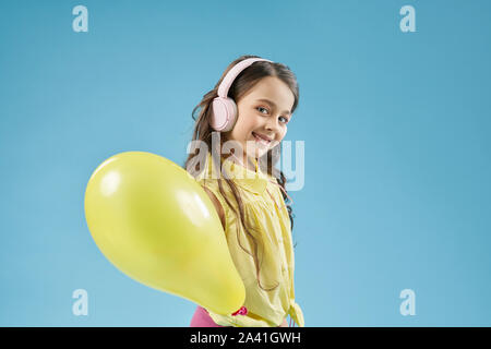Happy little girl wearing headphones keeping yellow balloon and looking at camera in studio. Cheerful child playing and having fun. Positive model posing on blue isolated background. Stock Photo