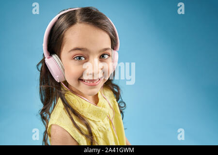 Positive child wearing pink earphones looking at camera and laughing on blue isolated background. Pretty little girl in yellow shirt listening music and enjoying modern device. Concept of technology. Stock Photo