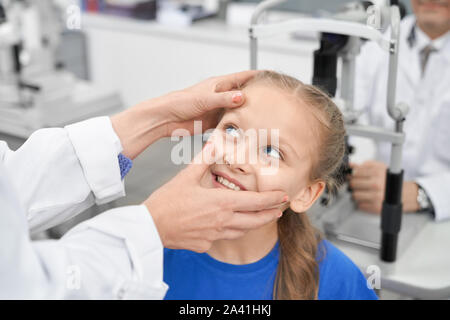 View from side of nice female oculist checking eye pupil of smiling girl in laboratory. Cheerful patient testing vision in clinic and choosing glasses or lenses. Concept of examination.
