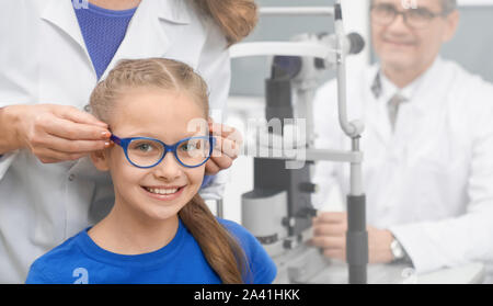 Front view of happy girl wearing new glasses in optical store. Pretty girl looking at camera and smiling while doctor offering glasses for improving vision. Concept of eye care and health. Stock Photo