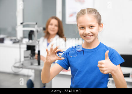 Front view of pretty girl in blue shirt showing sign ok and buying new glasses for vision. Young client looking at camera and choosing eyewear in optical shop. Concept of ophthalmology. Stock Photo