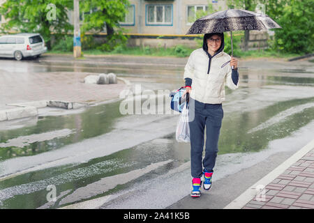 Young adult woman wearing casual sport clothes crossing road holding umbrella in hand during rain in autumn day. Girl walking on city street in autumn Stock Photo