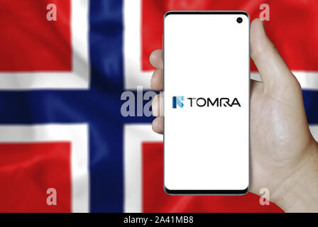 A man holds a phone displaying the logo of company Tomra Systems listed on Oslo Stock Exchange. Norwegian flag background. Credit: PIXDUCE Stock Photo