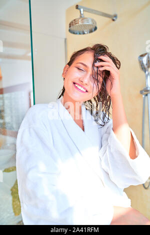 Photo of happy brunette with closed eyes with wet hair in white bathrobe standing in bath in apartment Stock Photo