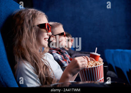 Side view of happy children wearing 3d glasses watching funny film in cinema. Excited friends eating popcorn, drinking aerated sweet water and having fun together. Concept of enjoyment and leisure. Stock Photo