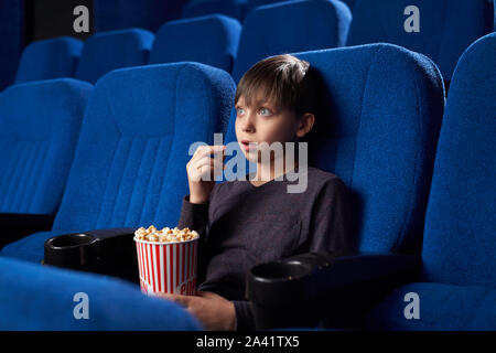 View from side of excited boy with big eyes and opened mouth watching amazing film in cinema. Shocked male teen eating popcorn, looking at screen and watching horror. Concept of leisure and fun.