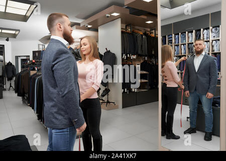 Side view of bearded male client looking at mirror and choosing new smart suit. Young man trying on fashionable jacket while woman measuring length of sleeve in boutique. Concept of clothes. Stock Photo