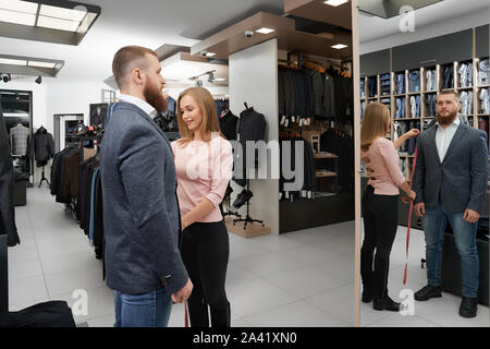View form side of stylish man wearing new grey jacket in boutique. Bearded man wearing smart suit standing in front of mirror and looking at reflection. Female assistant measuring length of sleeve. Stock Photo