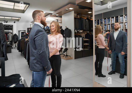 Attractive female tailor measuring length of sleeve of jacket in boutique. Young bearded male client trying on clothes, looking at mirror and choosing casual smart suit in shop. Concept of fitting. Stock Photo