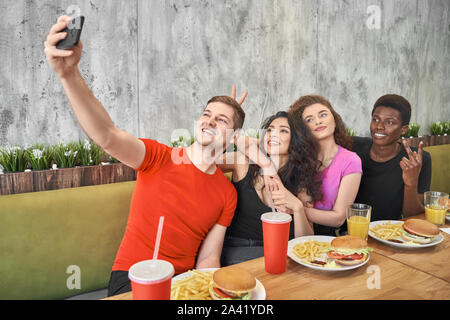 Friendly company doing selfie together in cafe while having lunch. Happy men and women looking at camera, posing and laughing in pizzeria. Cheerful students enjoying tasty fast food. Stock Photo