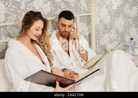 Newlyweds in hotel white coats woke up, look at the menu and make a phone order for Breakfast delivered to the room. Stock Photo
