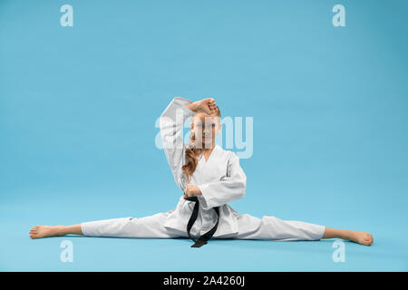 Confident girl in white kimono with black belt sitting on cross twine and looking at camera. Child training stretching and posing on blue background. Stock Photo