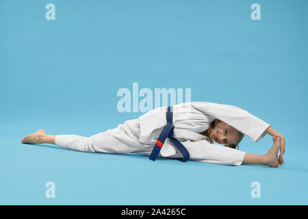 Positive karate girl sitting on longitudinal twine and reaching foot. Junior in white kimono and blue belt posing on light blue background in studio, looking at camera. Stock Photo