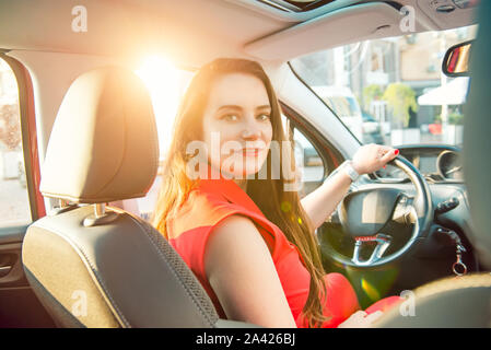 Back view Portrait of business lady, caucasian young woman driver looking at camera and smiling over her shoulder while driving a car. Sunset back lig Stock Photo