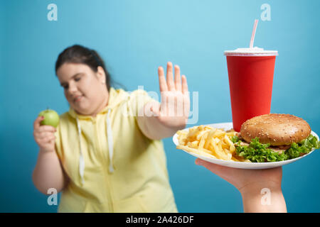 Portrait of chunky young lady in yellow sweater with closed eyes pushing out white plate with french fries, high calories burger and sparkling water in red cup. Concept of refusing harmful fast food Stock Photo