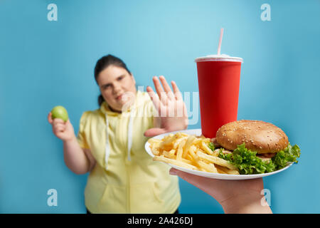 Young fat pretty brunette female in yellow sweater keeping fresh green apple in one hand and showing by another hand that she refuse to eat fastfood on white plate. Concept of weight loss Stock Photo