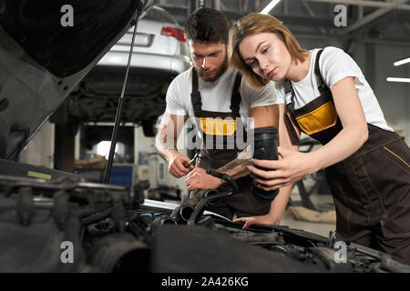 Side view of male and female mechanics repairing engine of automobile in auto service. Specialist in white shirt and coverall fixing car and changing tube in workshop. Concept of maintenance. Stock Photo