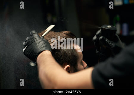 Crop of hairdresser's hands in black gloves combing clean male client hair with hairbrush and sprinkle it with water.Handsome man getting new hairstyle at modern and professional salon Stock Photo